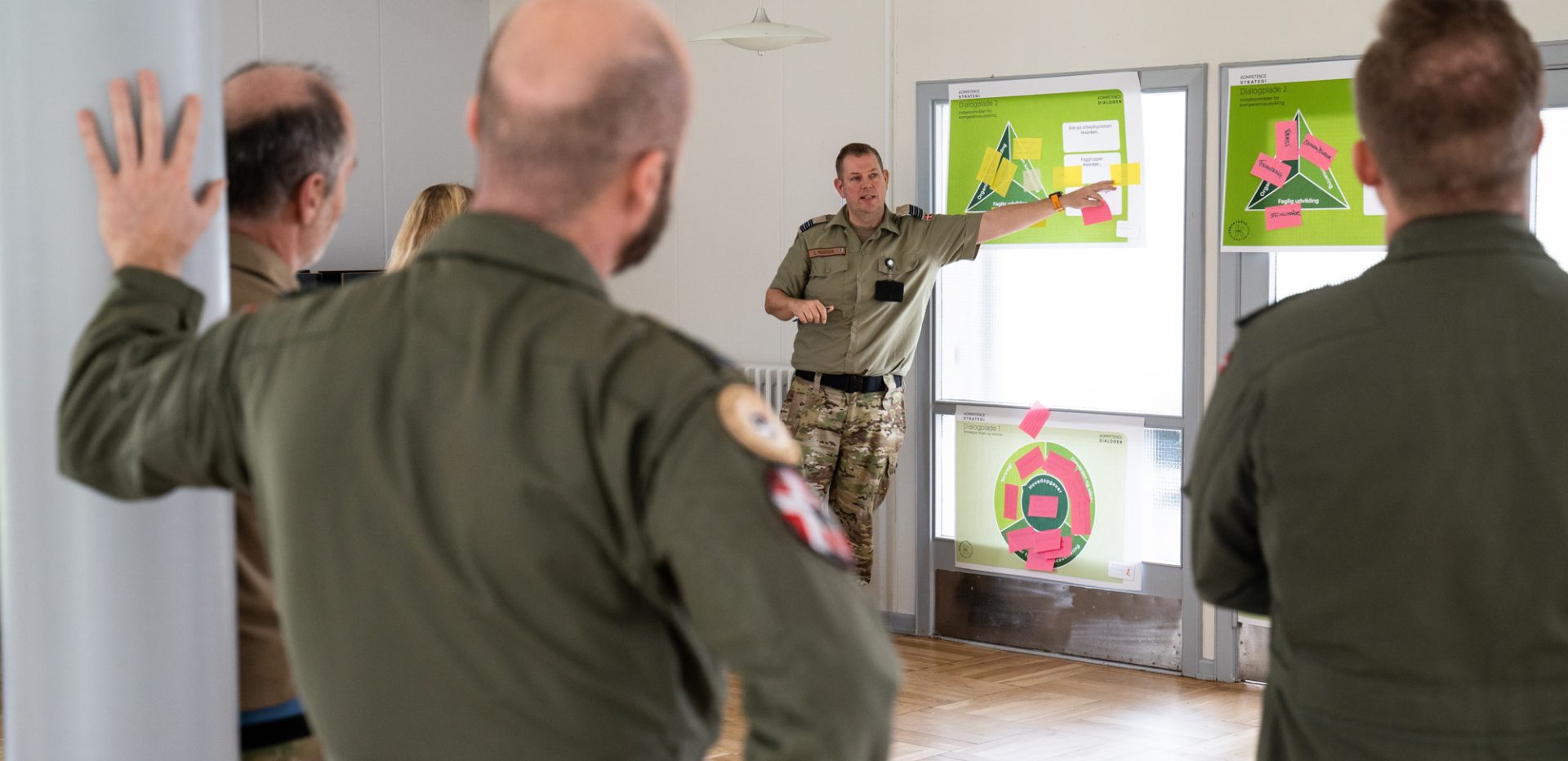 En soldat i uniform holder oplæg og peger på dialogplade fra Kompetencesekretariatet. To pilotter ser på oplæg.