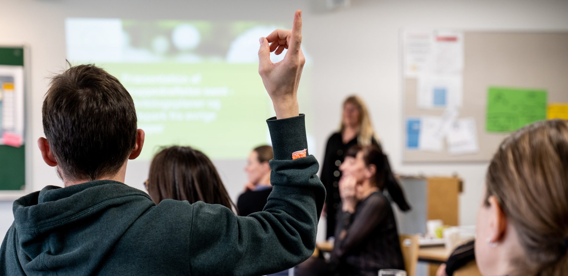 Farvefoto af person der rækker hånden op i noget der ligner et skolelokale